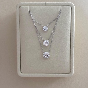 May include: Three silver necklaces with round, clear gemstones are displayed on a light beige background. The necklaces are layered, with each gemstone at a different level. The gemstones are the focal point of the jewelry.