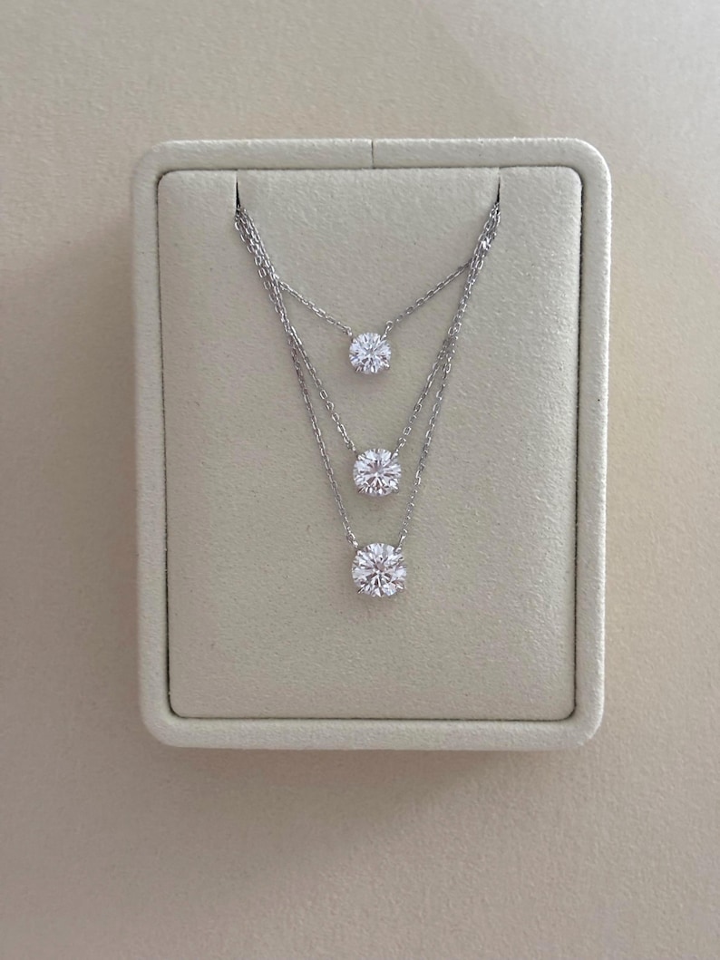May include: Three silver necklaces with round, clear gemstones are displayed on a light beige background. The necklaces are layered, with each gemstone at a different level. The gemstones are the focal point of the jewelry.
