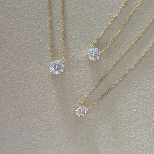 May include: Three gold necklaces with round, clear gemstones. Each necklace features a delicate gold chain and a single, sparkling stone. The necklaces are displayed on a neutral, textured surface, showcasing the jewelry's simple elegance.