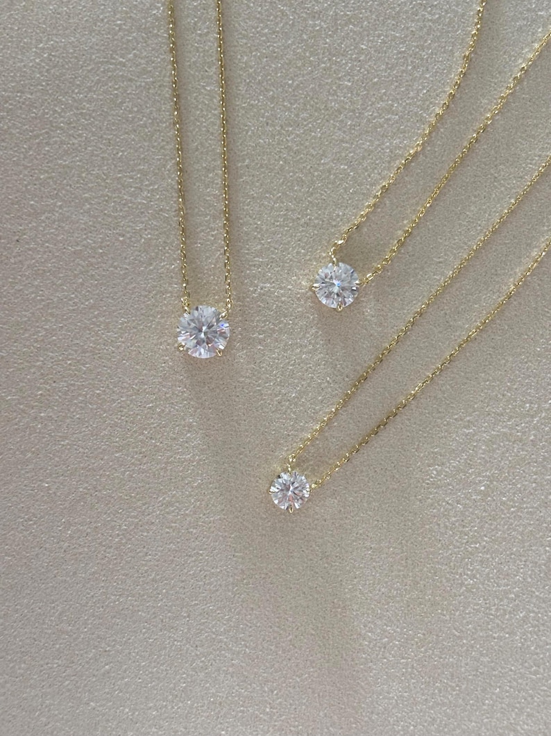 May include: Three gold necklaces with round, clear gemstones. Each necklace features a delicate gold chain and a single, sparkling stone. The necklaces are displayed on a neutral, textured surface, showcasing the jewelry's simple elegance.