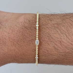 Puede incluir: Una pulsera de cuentas doradas que presenta una serie de pequeñas cuentas metálicas redondas intercaladas con varias piedras pequeñas, claras y de corte redondo, y una piedra central más grande de corte ovalado en una cadena fina.