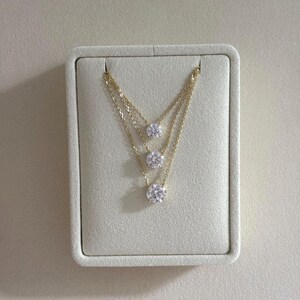 May include: Three-strand gold necklaces with round, clear gemstones. The necklaces are displayed on a light beige, rectangular jewelry display. The gemstones are evenly spaced along the gold chains.