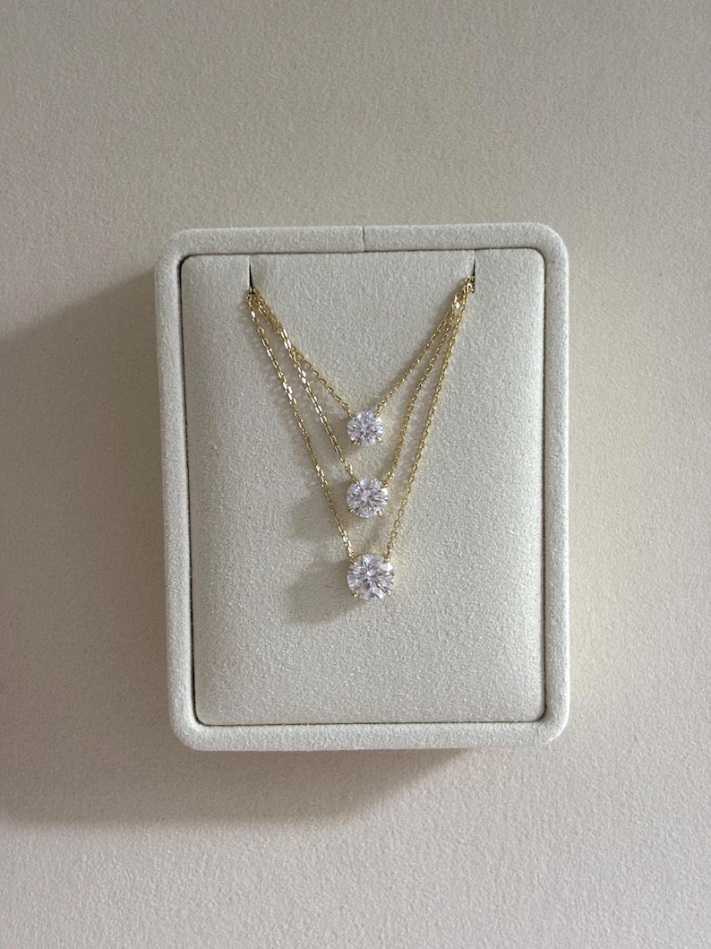 May include: Three-strand gold necklaces with round, clear gemstones. The necklaces are displayed on a light beige, rectangular jewelry display. The gemstones are evenly spaced along the gold chains.