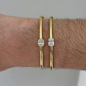 May include: Two gold-colored bangles with clear, sparkling gemstones. One bangle features an oval-shaped stone, while the other has a square-cut stone. The bracelets are worn on a wrist, showcasing their elegant design and shine.