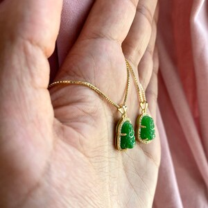May include: A gold chain necklace with two green jade pendants featuring a carved Buddha face. The pendants are set in a gold frame with small clear stones.