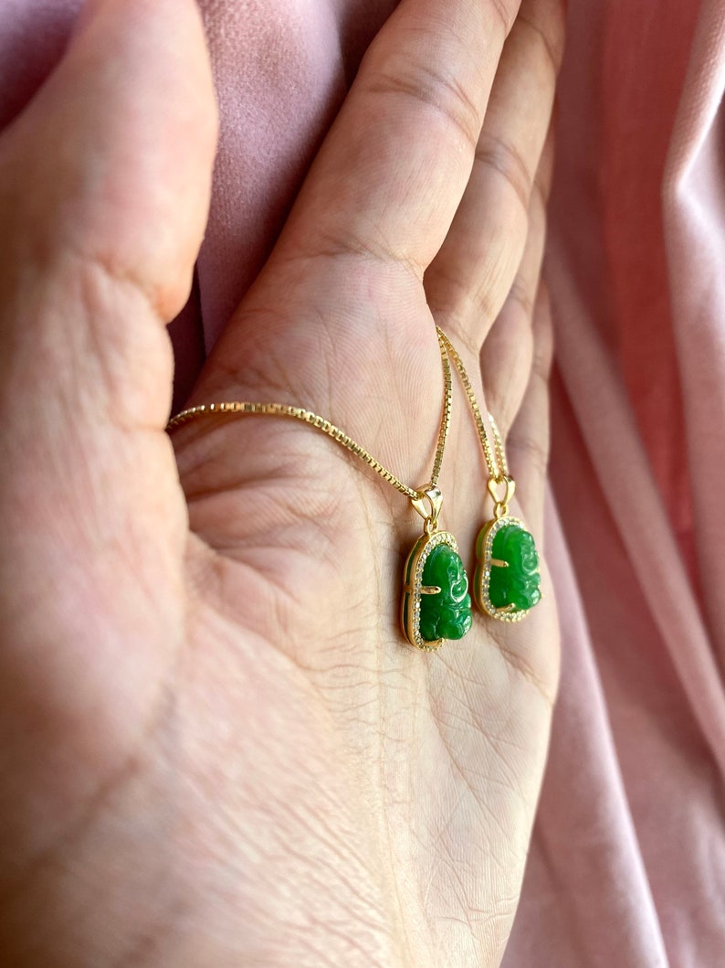 May include: A gold chain necklace with two green jade pendants featuring a carved Buddha face. The pendants are set in a gold frame with small clear stones.