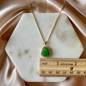May include: A gold chain necklace with a green jade Buddha pendant. The pendant is set with small clear stones. The necklace is measured against a ruler, showing it is just over 1 inch long.