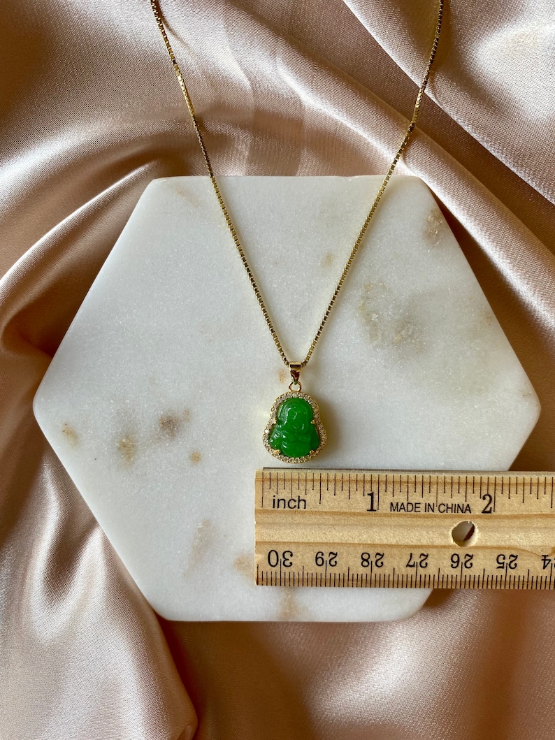 May include: A gold chain necklace with a green jade Buddha pendant. The pendant is set with small clear stones. The necklace is measured against a ruler, showing it is just over 1 inch long.