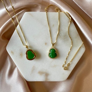 May include: Two gold chains with green jade pendants in the shape of a Buddha. The chains are on a white marble surface.