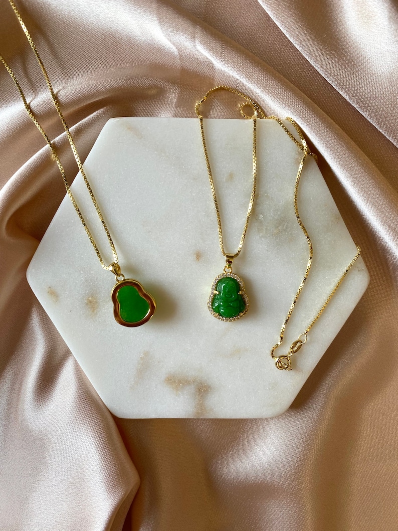 May include: Two gold chains with green jade pendants in the shape of a Buddha. The chains are on a white marble surface.