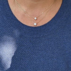May include: Two delicate gold necklaces, each featuring a single, round, clear gemstone pendant. The necklaces are layered, with one pendant slightly lower than the other. The person is wearing a blue sweater.