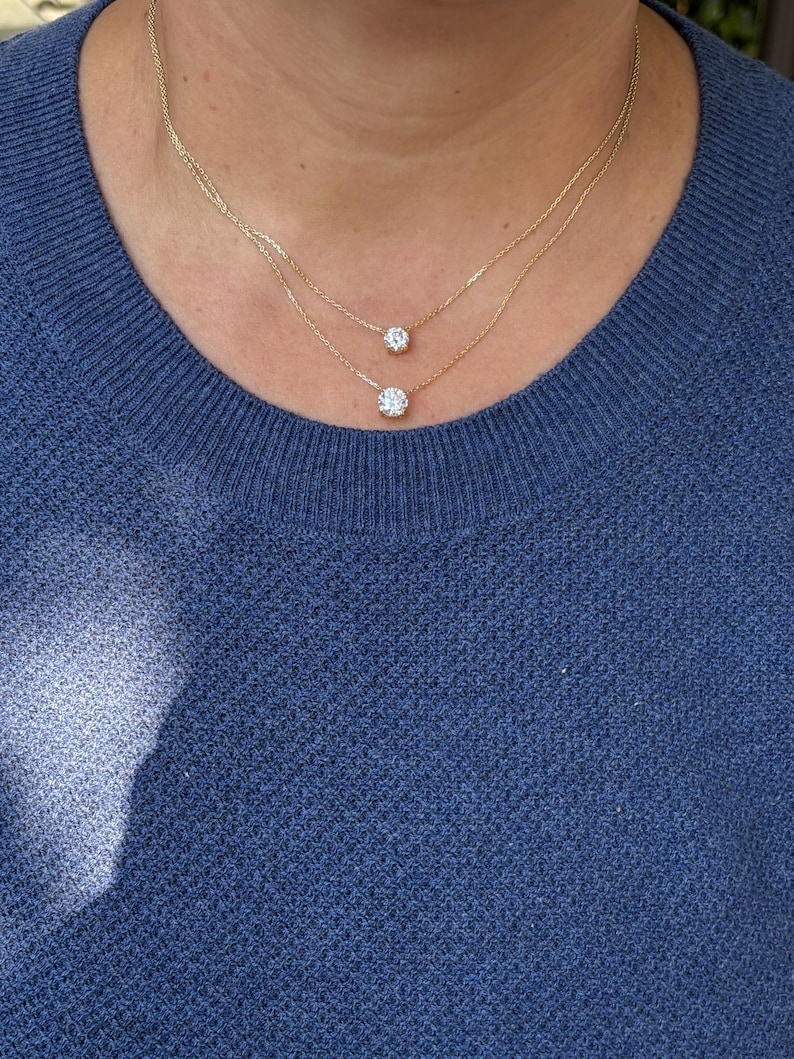 May include: Two delicate gold necklaces, each featuring a single, round, clear gemstone pendant. The necklaces are layered, with one pendant slightly lower than the other. The person is wearing a blue sweater.