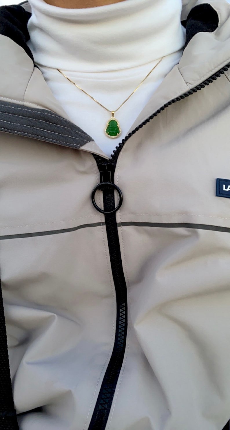 May include: A beige jacket with a black zipper and a green jade pendant necklace. The necklace has a gold chain and a small, green jade pendant in the shape of a Buddha.