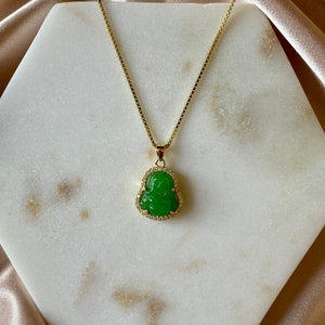May include: A gold chain necklace with a green jade pendant featuring a carved Buddha figure. The pendant is surrounded by small clear rhinestones.