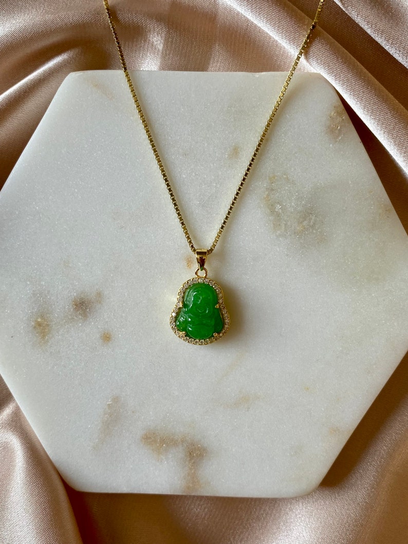 May include: A gold chain necklace with a green jade pendant featuring a carved Buddha figure. The pendant is surrounded by small clear rhinestones.