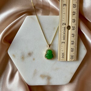 May include: A gold chain necklace with a green jade Buddha pendant. The pendant is set with small clear stones around the edge. The necklace is displayed on a white hexagonal tile.