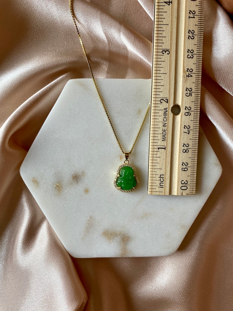 May include: A gold chain necklace with a green jade Buddha pendant. The pendant is set with small clear stones around the edge. The necklace is displayed on a white hexagonal tile.