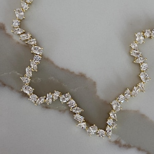 Può includere: Una collana dorata con una serie di pietre preziose chiare di forma irregolare. Le pietre sono disposte a zig-zag, creando un design scintillante ed elegante. La collana è esposta su una superficie chiara.