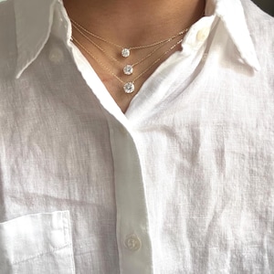 May include: Three gold necklaces with round, clear gemstones of varying sizes. The necklaces are layered, creating a cascading effect. The gemstones are set in a simple, classic style. The necklaces are worn over a white linen shirt.