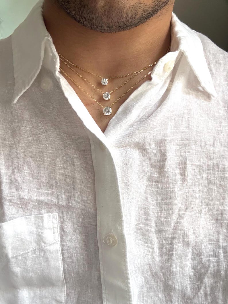 May include: Three gold necklaces with round, clear gemstones of varying sizes. The necklaces are layered, creating a cascading effect. The gemstones are set in a simple, classic style. The necklaces are worn over a white linen shirt.