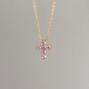Pode incluir: Um colar dourado com um pendente de cruz adornado com pedras preciosas rosa. A corrente e o pendente são exibidos sobre um fundo neutro, realçando o design e a cor da joia.