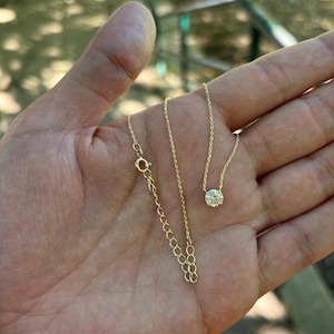 May include: A delicate gold necklace with a single, round, clear gemstone pendant. The necklace is displayed on an open hand, showcasing the fine chain and clasp. The background is blurred, suggesting an outdoor setting.