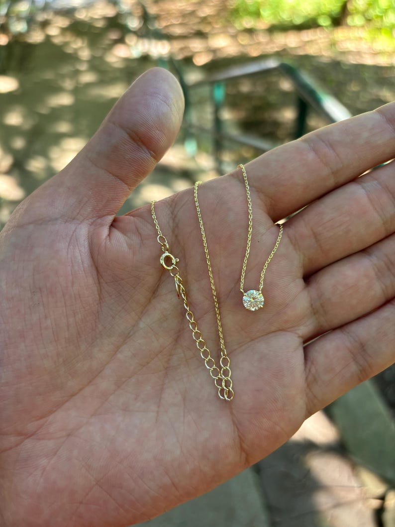 May include: A delicate gold necklace with a single, round, clear gemstone pendant. The necklace is displayed on an open hand, showcasing the fine chain and clasp. The background is blurred, suggesting an outdoor setting.