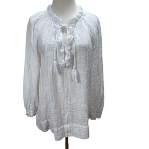 May include: White long-sleeve blouse with a textured weave. The blouse features a ruffled neckline, a tie closure at the chest, and long sleeves with ruffled cuffs. The fabric appears lightweight and semi-sheer, suitable for warmer weather.