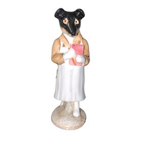 May include: A ceramic figurine of a dog with a black and white head, wearing a tan jacket over a white dress. The dog holds a pink rectangular object and a small orange pencil in its white-gloved hands, standing on a circular base.