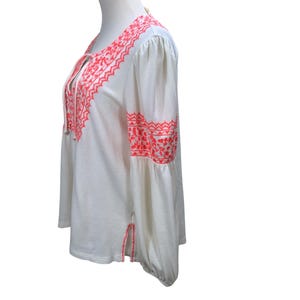 May include: White long-sleeve blouse with coral embroidery. The blouse features a V-neck with tie closure and decorative embroidery along the neckline, sleeves, and hem. The sleeves are long and flared, and the fabric has a subtle textured pattern.