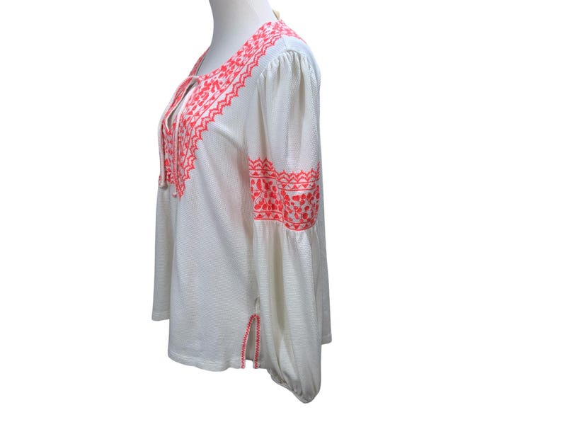 May include: White long-sleeve blouse with coral embroidery. The blouse features a V-neck with tie closure and decorative embroidery along the neckline, sleeves, and hem. The sleeves are long and flared, and the fabric has a subtle textured pattern.
