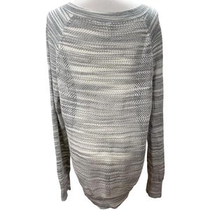 Puede incluir: Un su&eacute;ter de punto gris y blanco con cuello barco y mangas largas. El su&eacute;ter tiene un patr&oacute;n texturizado y se muestra en un maniqu&iacute; blanco. La prenda de punto es de color claro y neutro, adecuada para combinar.