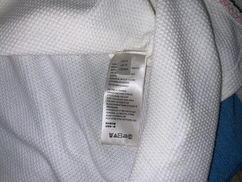 May include: Close-up of a white textured fabric with a tag attached. The tag includes washing instructions and size information. The fabric has a subtle, grid-like pattern, and the tag indicates a gentle wash cycle.