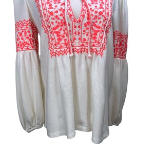 May include: Off-white long-sleeve top with vibrant coral embroidery. The top features a V-neckline with a tie, and the embroidery adorns the neckline and sleeves. The sleeves are long and gathered at the wrist.