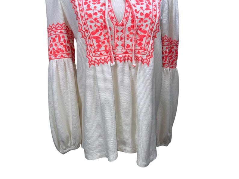 May include: Off-white long-sleeve top with vibrant coral embroidery. The top features a V-neckline with a tie, and the embroidery adorns the neckline and sleeves. The sleeves are long and gathered at the wrist.