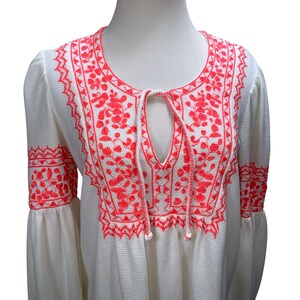 May include: White tunic top with bright coral embroidery. The embroidery features floral and geometric patterns around the neckline, keyhole, and sleeves. The top has long, bell sleeves and a textured fabric.