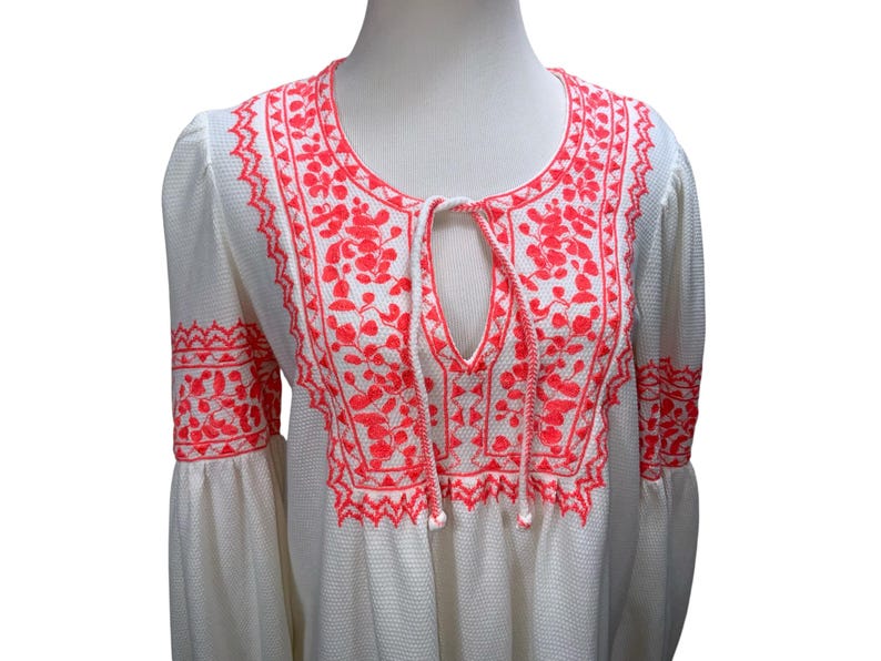 May include: White tunic top with bright coral embroidery. The embroidery features floral and geometric patterns around the neckline, keyhole, and sleeves. The top has long, bell sleeves and a textured fabric.