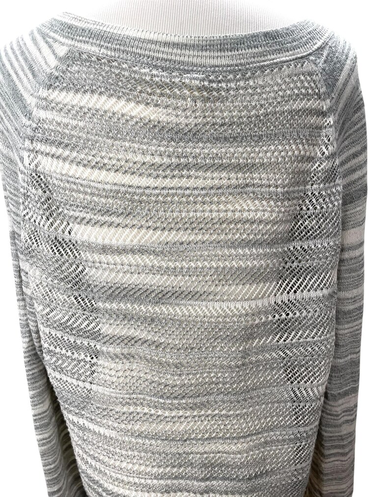 Puede incluir: Un su&eacute;ter a rayas grises y blancas con cuello barco. El su&eacute;ter tiene un dise&ntilde;o de punto texturizado con detalles calados en los laterales. Las mangas son largas y el estilo general es informal.