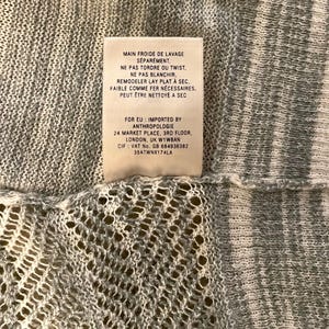 Puede incluir: Primer plano de una tela de punto gris claro con una etiqueta adjunta. La etiqueta tiene texto en franc&eacute;s, incluidas las instrucciones de lavado e informaci&oacute;n de importaci&oacute;n de Anthropologie, Londres.