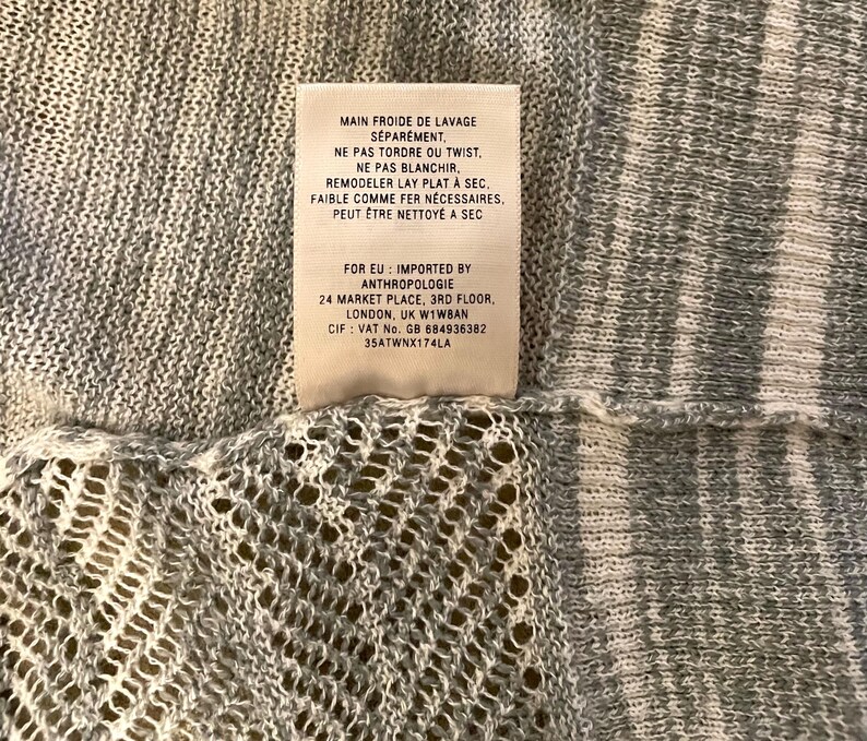 Puede incluir: Primer plano de una tela de punto gris claro con una etiqueta adjunta. La etiqueta tiene texto en franc&eacute;s, incluidas las instrucciones de lavado e informaci&oacute;n de importaci&oacute;n de Anthropologie, Londres.