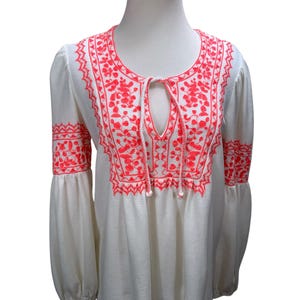 May include: White long-sleeve top with bright coral embroidery. The embroidery features floral and geometric patterns around the neckline, cuffs, and upper chest. The top has a keyhole neckline with a drawstring.