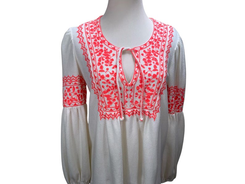 May include: White long-sleeve top with bright coral embroidery. The embroidery features floral and geometric patterns around the neckline, cuffs, and upper chest. The top has a keyhole neckline with a drawstring.