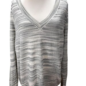 Puede incluir: Un su&eacute;ter de cuello en V de manga larga con un patr&oacute;n de rayas horizontales grises y blancas. El su&eacute;ter se muestra en un maniqu&iacute;, mostrando su ajuste relajado y su textura suave. El escote y los pu&ntilde;os tienen un sutil detalle acanalado.