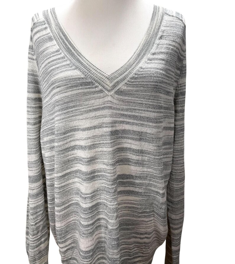 Puede incluir: Un su&eacute;ter de cuello en V de manga larga con un patr&oacute;n de rayas horizontales grises y blancas. El su&eacute;ter se muestra en un maniqu&iacute;, mostrando su ajuste relajado y su textura suave. El escote y los pu&ntilde;os tienen un sutil detalle acanalado.