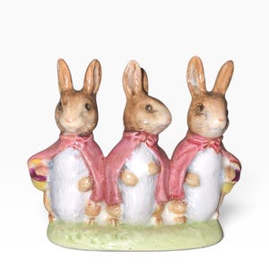 Könnte beinhalten: Eine Keramikfigur mit drei braun-weißen Kaninchen, die nebeneinander auf einem hellgrünen Sockel stehen. Jedes Kaninchen trägt einen am Hals gebundenen rosa Umhang. Die Kaninchen haben braune Ohren, weiße Bäuche und stehen in einer Reihe.