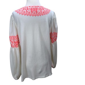 May include: White long-sleeve blouse with bright pink embroidery on the neckline and sleeves. The blouse has a textured fabric and a loose, flowing fit. The sleeves are long and gathered at the wrist, creating a bell shape.