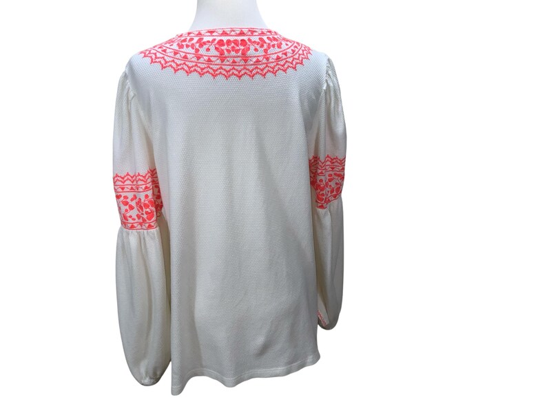 May include: White long-sleeve blouse with bright pink embroidery on the neckline and sleeves. The blouse has a textured fabric and a loose, flowing fit. The sleeves are long and gathered at the wrist, creating a bell shape.