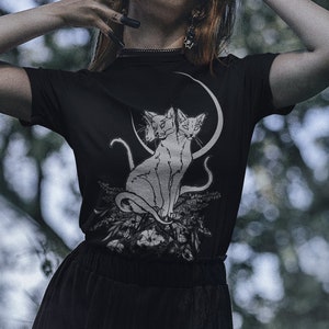 May include: A woman wearing a black t-shirt with a white graphic of two cats under a crescent moon. She is wearing a black skirt and has black nail polish.