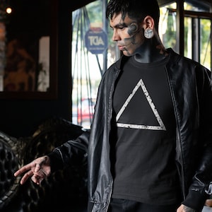 以下が含まれることがあります： 黒い革のジャケットを黒いTシャツの上に着ている人。Tシャツには白い三角形のグラフィックがプリントされています。三角形は、使い古した斑点のある質感です。