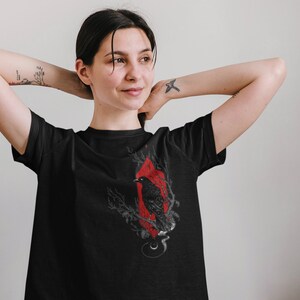May include: Black t-shirt with a graphic print of a raven in a red diamond shape with branches and a crescent moon. The raven is perched on a branch with its wings spread.
